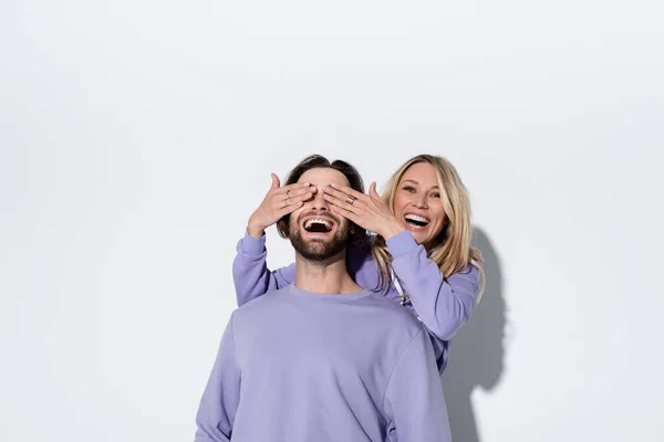 Happy blonde woman covering eyes of bearded man in purple sweatshirt on grey — Photo de stock