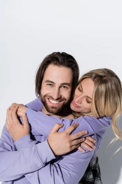 Happy blonde woman hugging smiling bearded man in purple sweatshirt isolated on grey - foto de stock