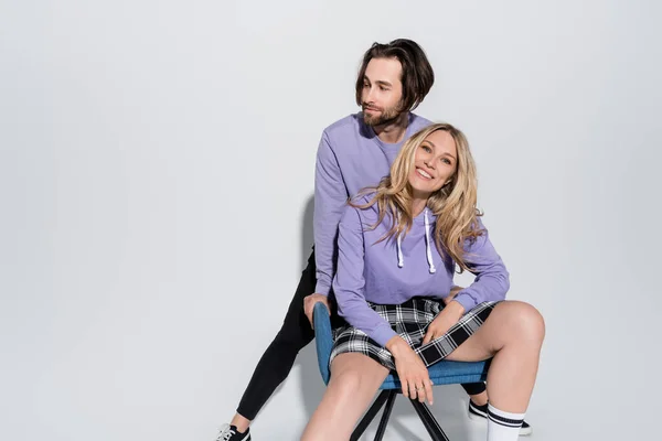 Cheerful man standing near happy blonde woman in tartan skirt sitting on blue armchair on grey — Foto stock