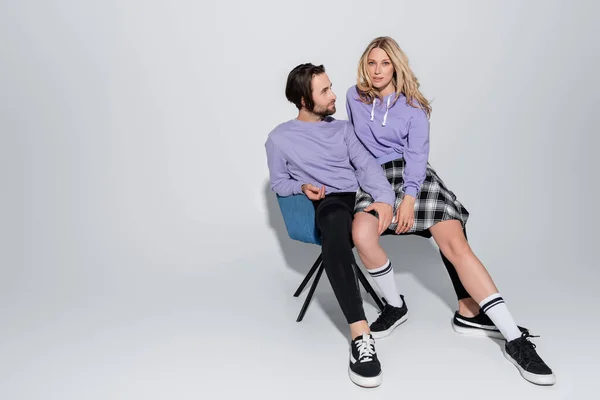 Full length of cheerful man and blonde woman in tartan skirt sitting on blue armchair on grey - foto de stock