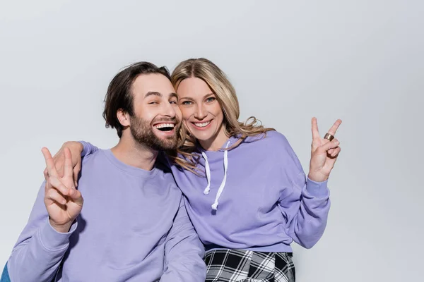 Happy couple in purple sweatshirts showing peace sign and smiling isolated on grey — Stockfoto