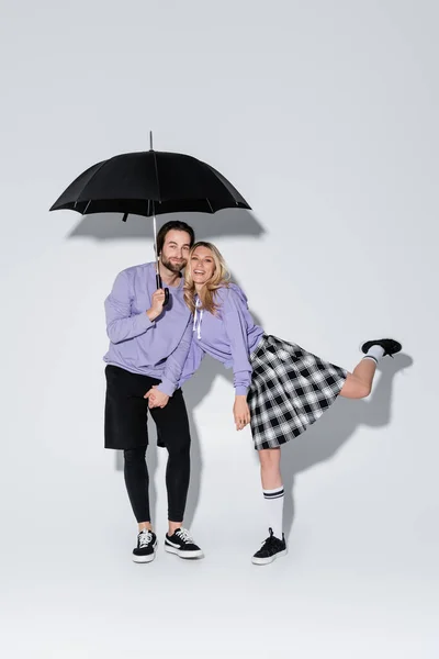 Full length of happy couple in purple sweatshirts standing under umbrella on grey — Fotografia de Stock