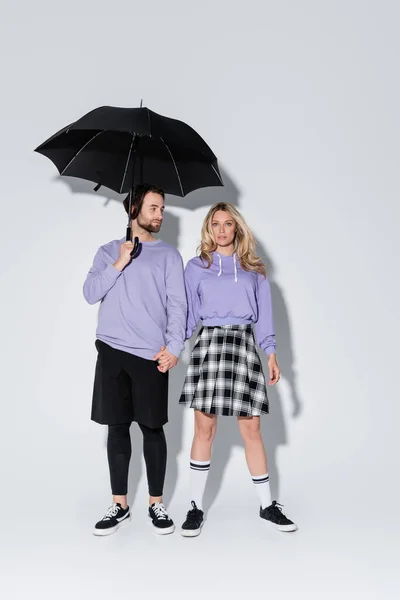 Full length of stylish couple in purple sweatshirts standing under black umbrella on grey — Fotografia de Stock