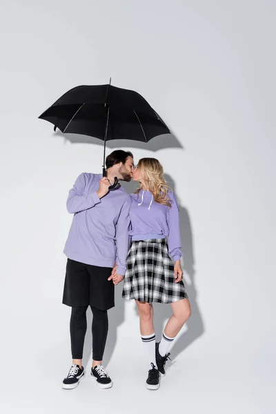 Full length of happy couple in purple sweatshirts kissing while standing under umbrella on grey — Stock Photo