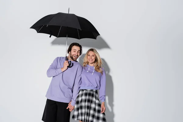 Happy couple in purple sweatshirts smiling while standing under umbrella isolated on grey — Fotografia de Stock