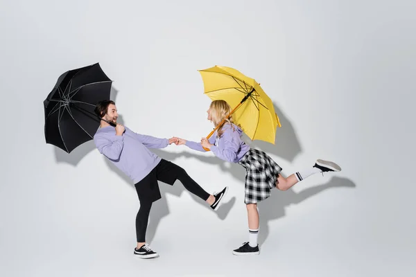 Full length of cheerful couple in purple sweatshirts holding hands while standing under umbrellas on grey — Stock Photo