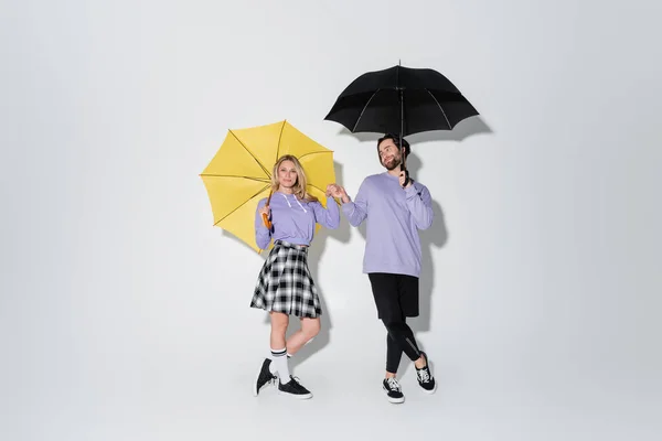 Full length of pleased couple in purple sweatshirts holding hands while standing under umbrellas on grey — Foto stock