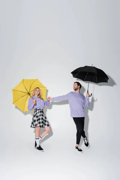 Full length of joyful couple in purple sweatshirts holding hands while standing under umbrellas on grey — Foto stock