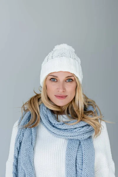 Портрет блондинки в зимней шляпе и вязаном шарфе, смотрящей на камеру, изолированную на сером — стоковое фото
