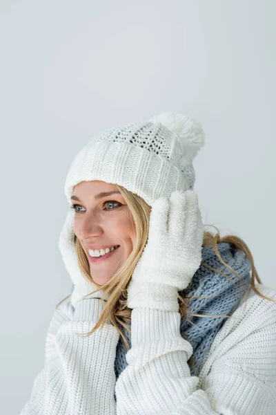 Portrait of blonde woman in winter hat and white gloves touching face and smiling isolated on grey — Photo de stock