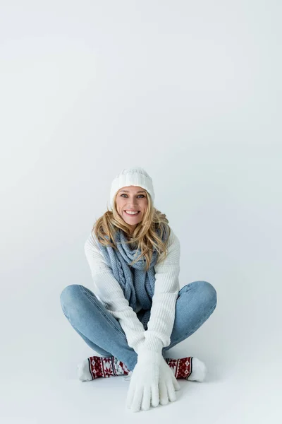 Happy blonde woman in winter hat and knitted scarf looking at camera while sitting on grey — Stock Photo