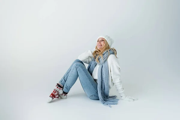 Joyful blonde woman in white knitted hat and winter outfit sitting on grey — Stockfoto