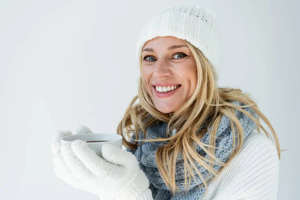 Portrait of happy woman in winter hat and white gloves holding cup of hot beverage isolated on grey — Foto stock