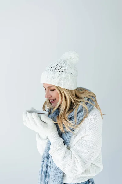 Portrait of cheerful woman in winter hat and white gloves holding cup of hot beverage isolated on grey — Stock Photo