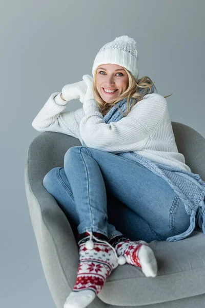 Cheerful blonde woman in winter outfit sitting in comfortable armchair isolated on grey - foto de stock