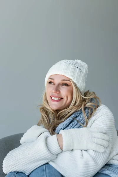 Dreamy blonde woman in winter outfit sitting in comfortable armchair and smiling isolated on grey — Fotografia de Stock