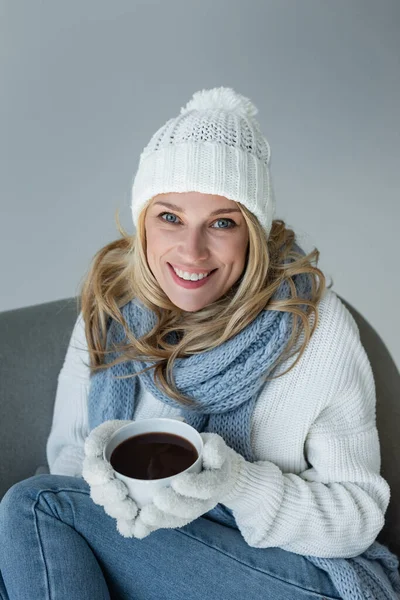 Happy blonde woman in winter outfit sitting in comfortable armchair and holding cup of coffee isolated on grey — Foto stock