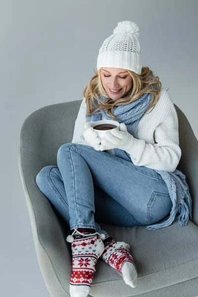 Cheerful blonde woman in winter outfit sitting in comfortable armchair and holding cup of coffee isolated on grey — Photo de stock