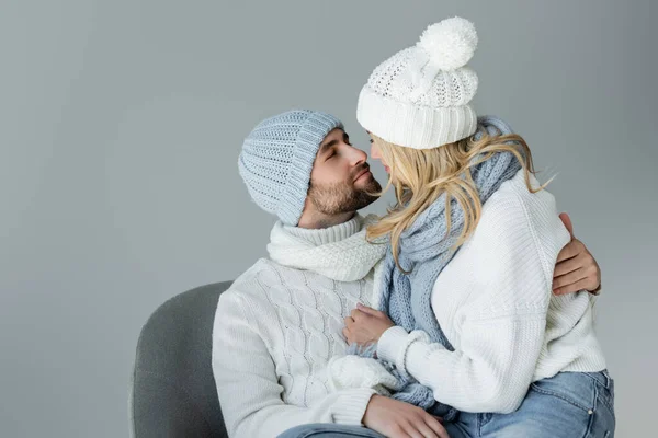 Blonde woman in winter hat and knitted scarf looking at bearded boyfriend in armchair isolated on grey — Stockfoto