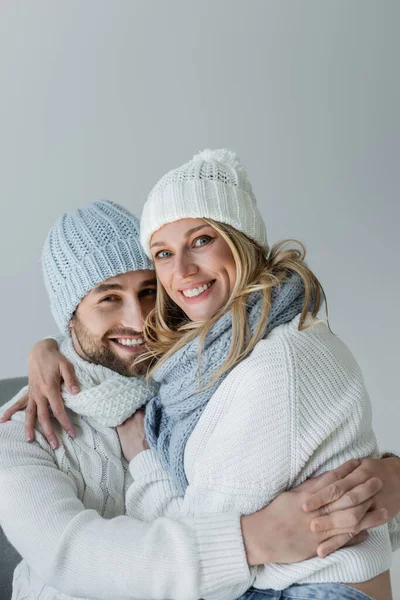 Happy blonde woman in knitted hat hugging with smiling boyfriend in winter outfit isolated on grey - foto de stock