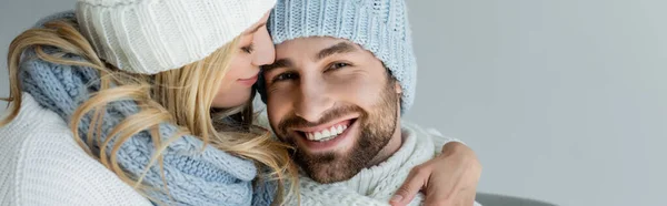 Блондинка в вязаной шляпе обнимает улыбающегося бойфренда в зимнем наряде, изолированном на сером, баннер — стоковое фото