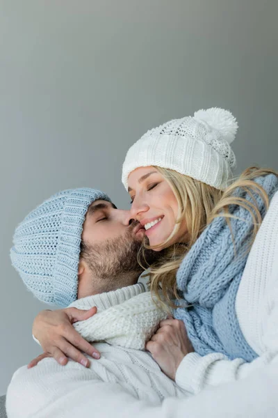 Bearded man kissing cheek of happy blonde woman in knitted hat isolated on grey — Photo de stock