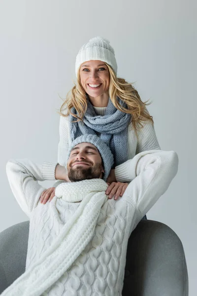 Happy blonde woman in knitted hat near smiling boyfriend in winter outfit sitting in armchair isolated on grey - foto de stock