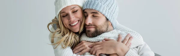 Happy blonde woman in knitted hat hugging with smiling boyfriend in winter outfit isolated on grey, banner — Fotografia de Stock