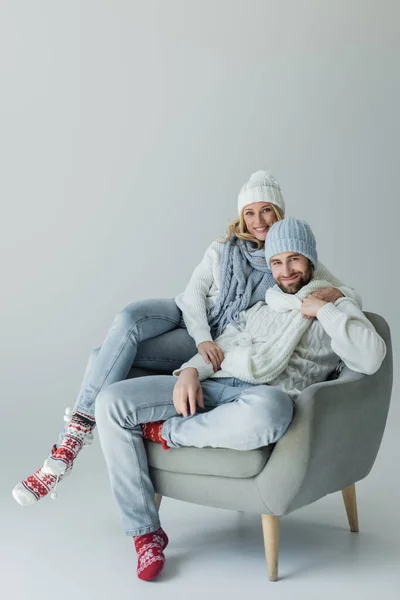 Happy blonde woman in knitted hat hugging with smiling boyfriend in winter outfit while sitting in armchair on grey — Foto stock