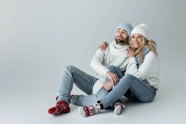 Full length of happy blonde woman in knitted hat hugging with smiling boyfriend in winter outfit while sitting on grey - foto de stock