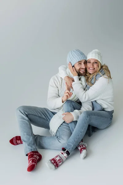 Full length of happy blonde woman in knitted hat embracing cheerful boyfriend in winter outfit while sitting on grey - foto de stock