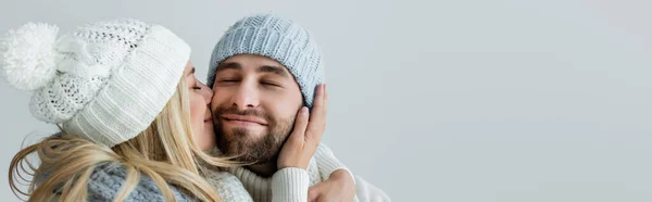 Happy blonde woman in knitted hat kissing cheek of smiling boyfriend isolated on grey, banner — Stockfoto