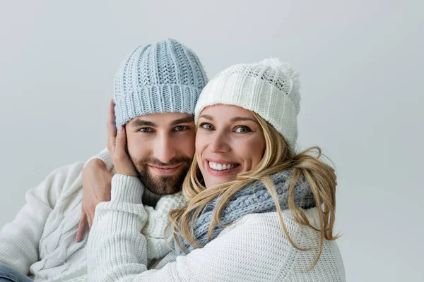 Happy blonde woman in knitted hat hugging pleased and bearded boyfriend isolated on grey — Stockfoto