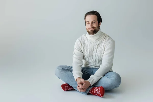 Full length of bearded man in jeans and knitted sweater sitting in red socks with ornament on grey — Fotografia de Stock