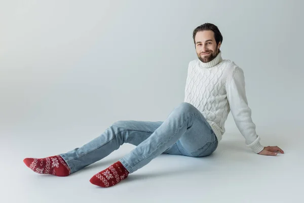 Full length of bearded man in jeans and white sweater sitting in red socks with ornament on grey - foto de stock