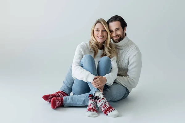 Full length of smiling couple in winter outfits and red socks with ornament sitting on grey — Fotografia de Stock