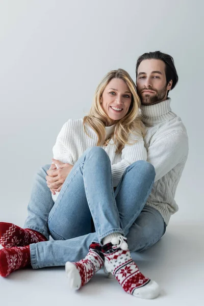 Full length of cheerful couple in winter outfits and red socks with ornament sitting on grey - foto de stock
