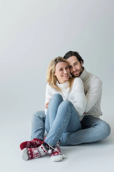 Full length of happy couple in winter outfits and red socks with ornament sitting on grey — Stockfoto