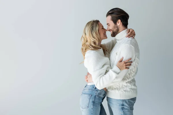 Side view of cheerful couple in knitted white sweaters and jeans smiling while hugging isolated on grey — Stockfoto