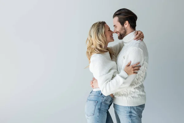 Side view of cheerful couple in knitted white sweaters and jeans smiling while hugging isolated on grey — Stock Photo