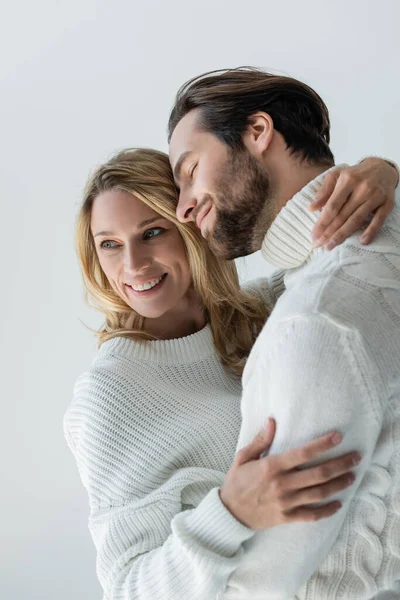 Cheerful couple in knitted white sweaters and jeans smiling while hugging isolated on grey — Photo de stock