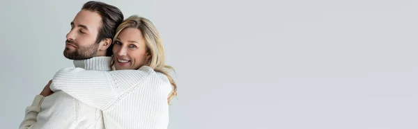 Cheerful blonde woman in white knitted sweater hugging bearded man isolated on grey, banner — Foto stock