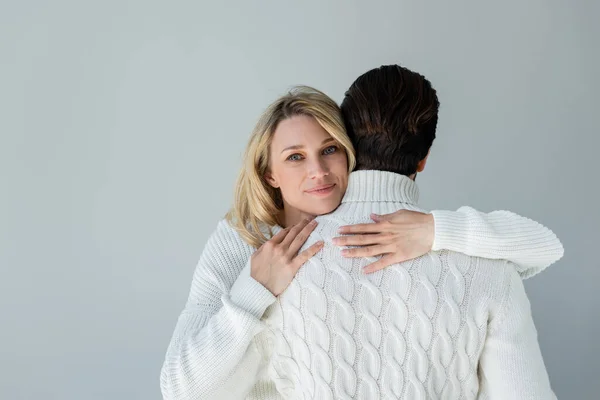 Blonde woman in white sweater hugging back of boyfriend and smiling isolated on grey — Photo de stock