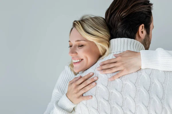 Cheerful blonde woman in white sweater hugging back of boyfriend isolated on grey — Stockfoto