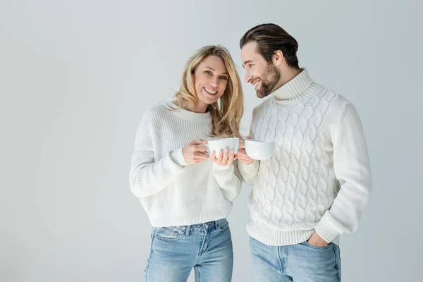 Happy man and cheerful woman in white knitted sweaters holding cups of coffee isolated on grey — Photo de stock