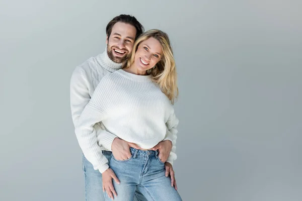 Счастливая пара в белых свитерах и джинсах в джинсах, позирующих изолированно на сером — стоковое фото