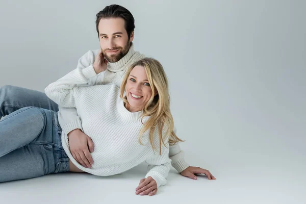 Cheerful man and woman in white knitted sweaters looking at camera on grey — Fotografia de Stock