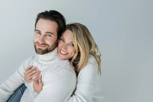 Cheerful woman hugging bearded man in white sweater and looking at camera isolated on grey - foto de stock