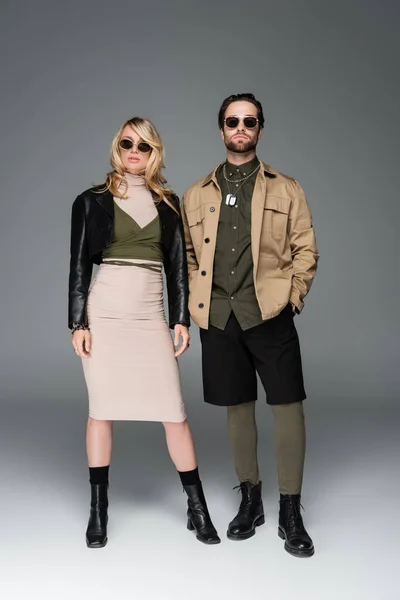 Full length of stylish couple in fashionable outfits and sunglasses posing together on grey — Stockfoto