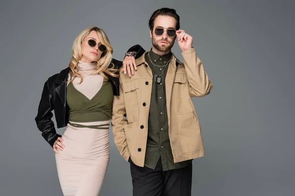Stylish man adjusting sunglasses near blonde woman in leather jacket isolated on grey - foto de stock